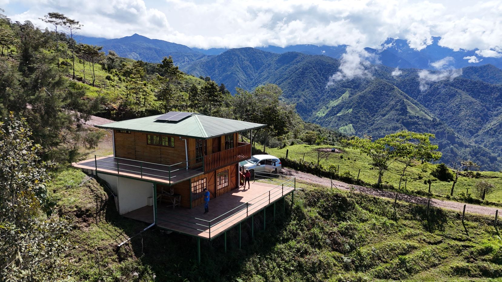 Wooden hillside cabin with a wraparound deck, solar panel on the green roof, and a car parked nearby against a backdrop of lush, misty mountains.