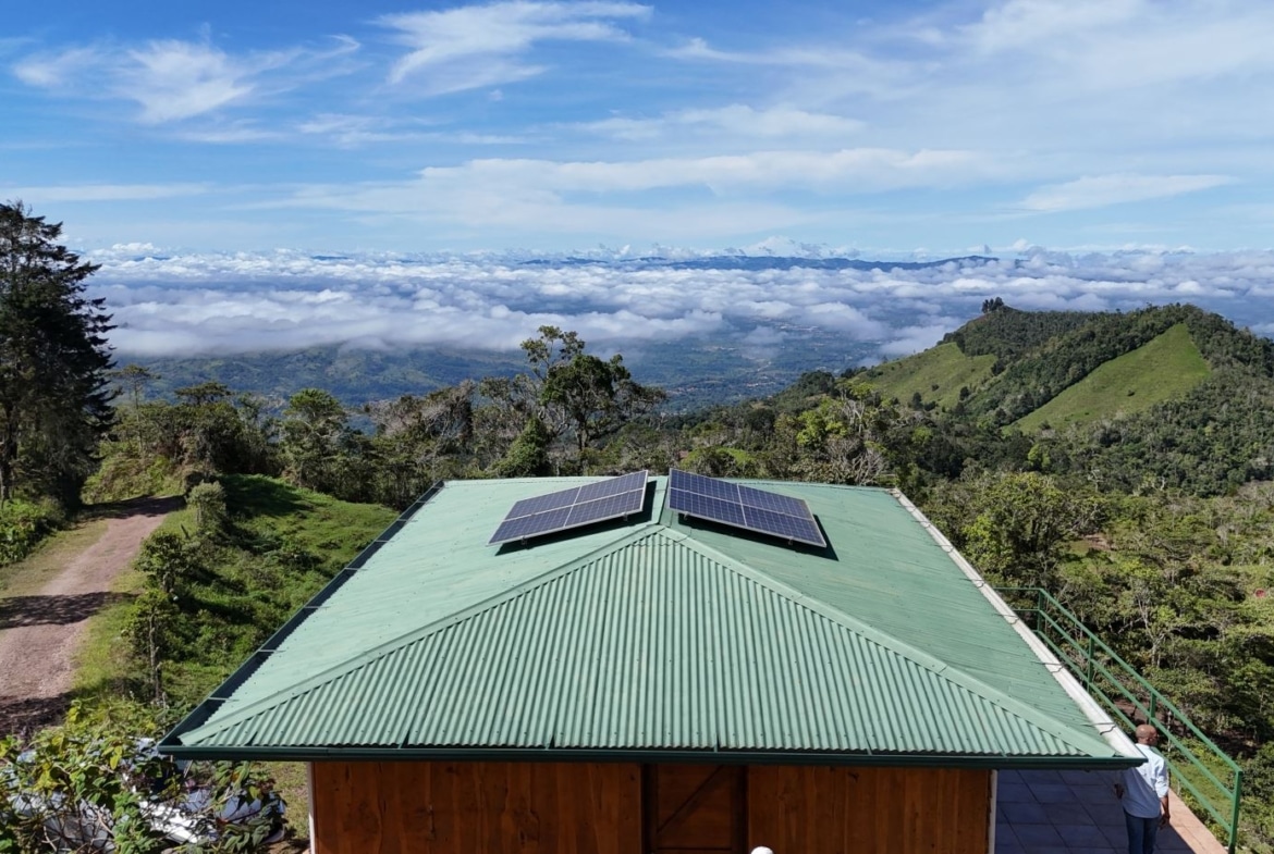 Green metal-roof building with two solar panels, set in a hilly landscape under a blue sky and fluffy clouds.