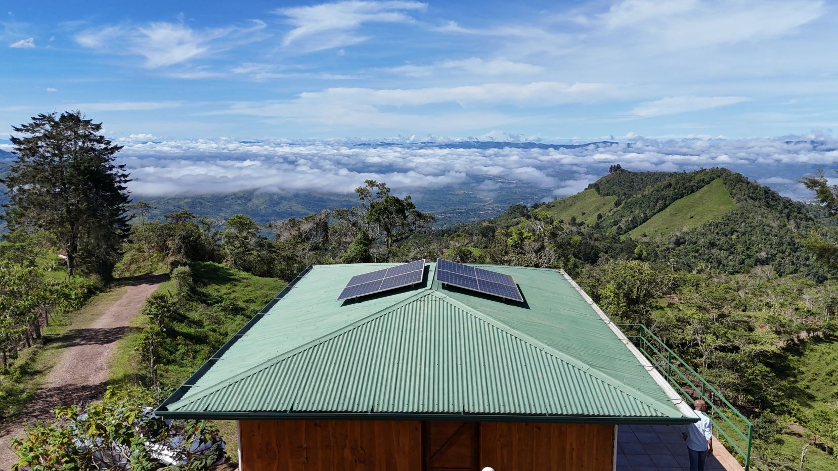 Green metal-roof building with two solar panels, set in a hilly landscape under a blue sky and fluffy clouds.