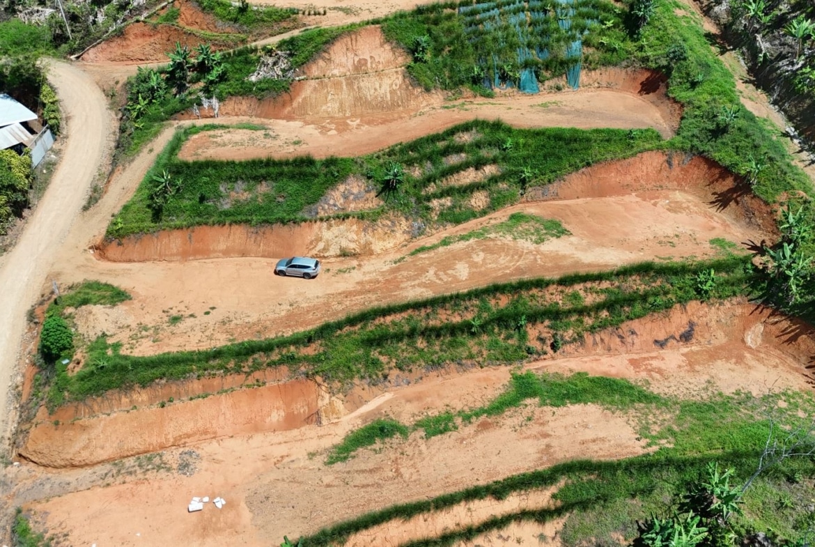 Aerial view of terraced red-sand hills with green vegetation; a silver SUV is parked on one terrace beside a winding dirt road.