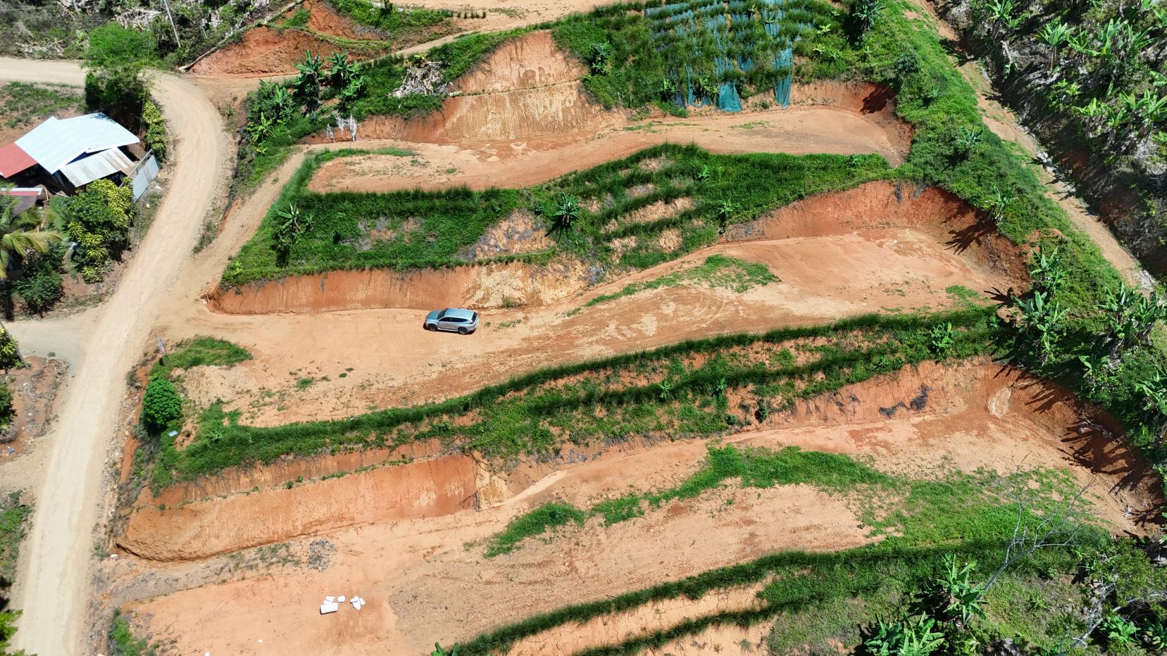 Aerial view of terraced red-sand hills with green vegetation; a silver SUV is parked on one terrace beside a winding dirt road.
