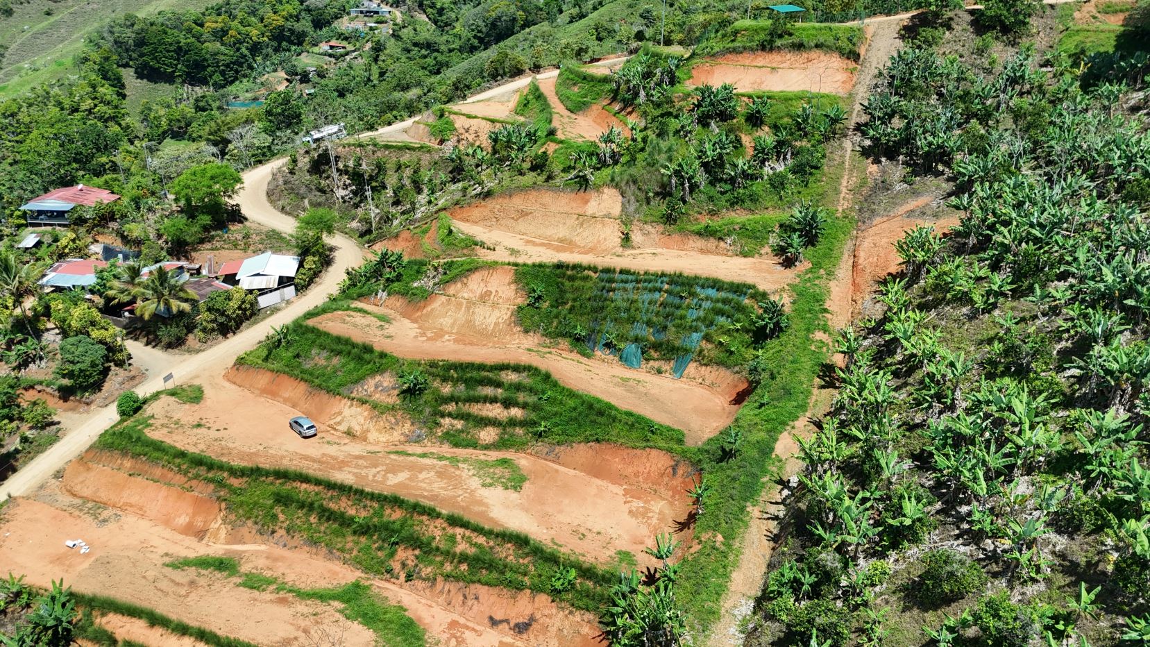 Aerial view of terraced hillside farmland with orange soil, banana trees, and a winding dirt road through a rural village on the left.