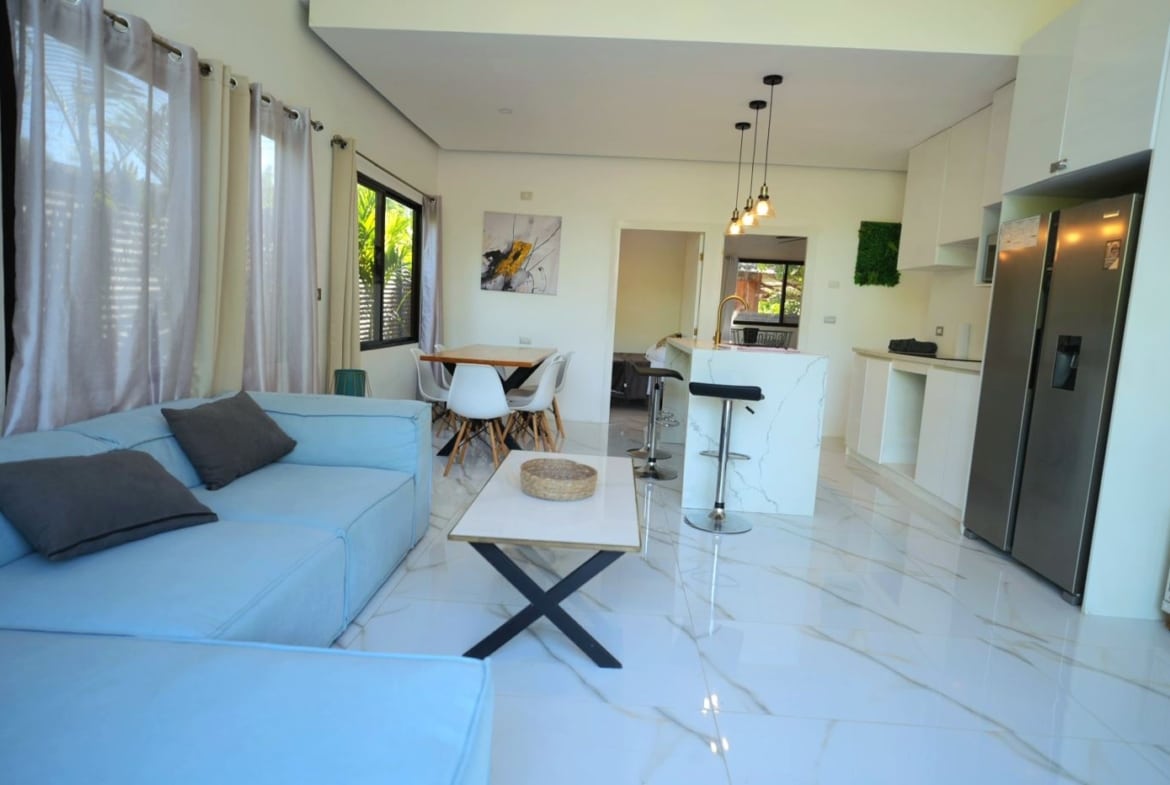 Bright open-plan living area with a light blue sofa, white furniture, and a marble-topped kitchen island.