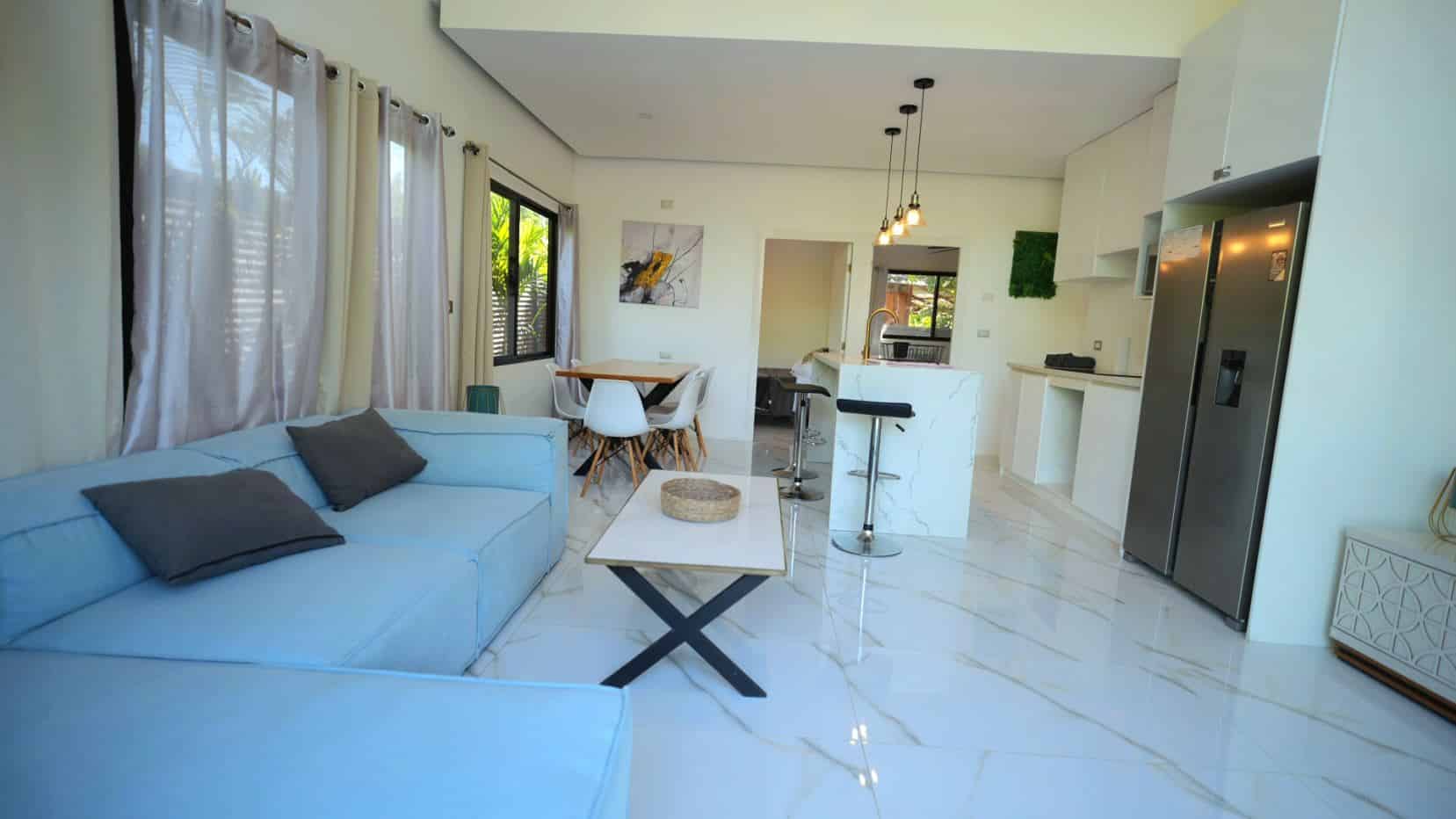 Bright open-plan living area with a light blue sofa, white furniture, and a marble-topped kitchen island.