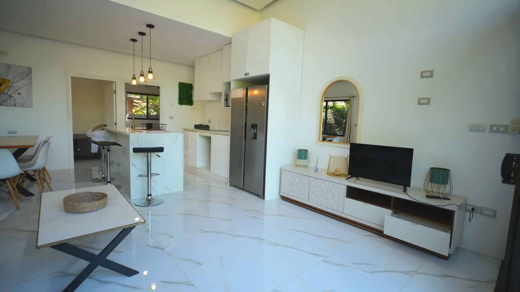 Open-plan living area with white marble floors, a marble-topped kitchen island, bar stools, and a stainless steel fridge.