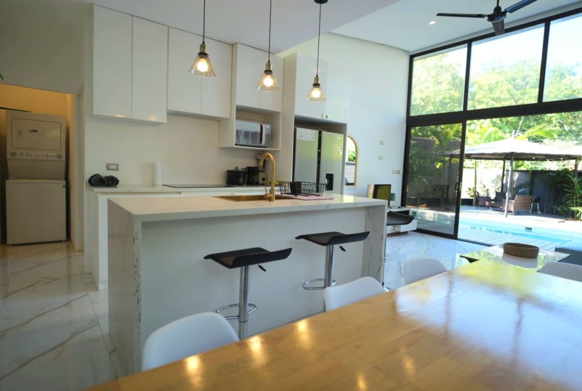 Open-plan kitchen with a marble island, pendant lights, white cabinets, and a dining area adjacent to glass doors overlooking a pool.