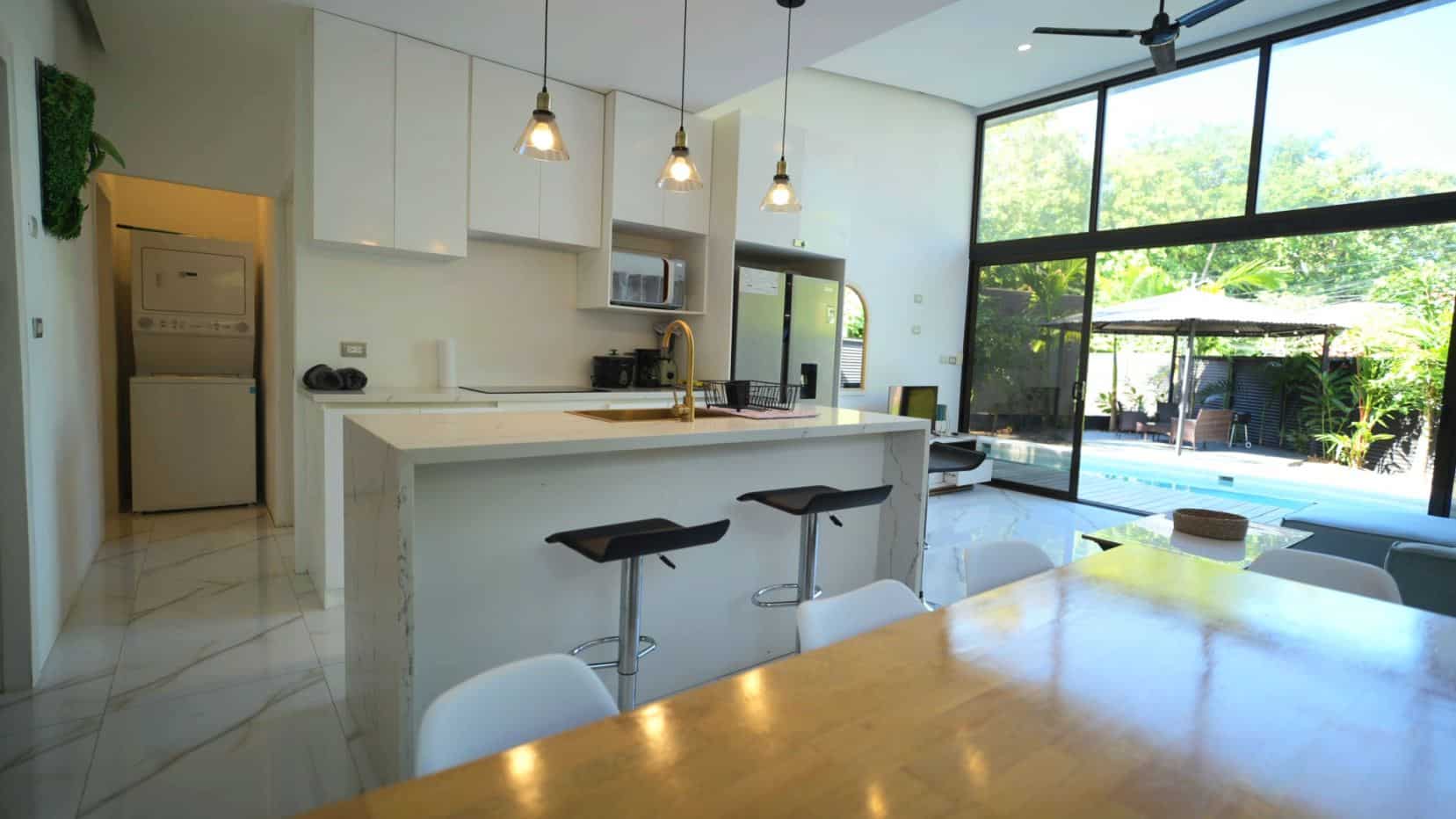 Open-plan kitchen with a marble island, pendant lights, white cabinets, and a dining area adjacent to glass doors overlooking a pool.