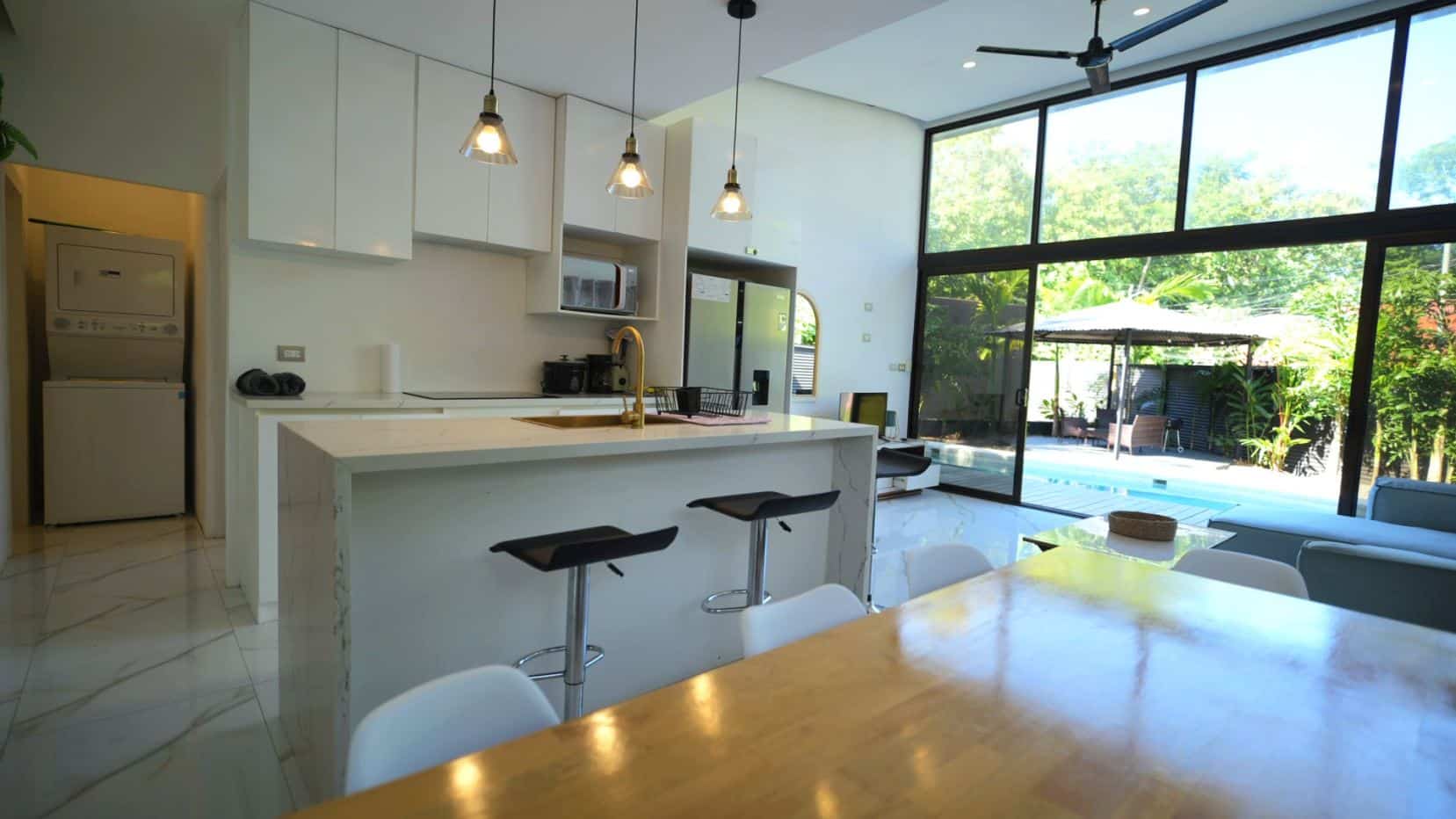 Open-concept kitchen with white cabinets and a marble island, pendant lights, and floor-to-ceiling windows overlooking a sunny garden.
