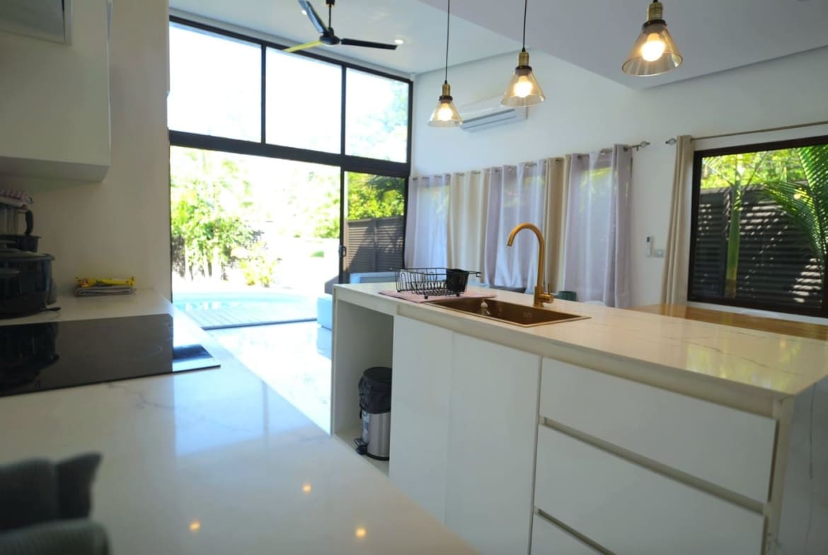 Bright, modern kitchen with white cabinetry and marble island, featuring a gold faucet and large windows overlooking a sunny outdoor space.
