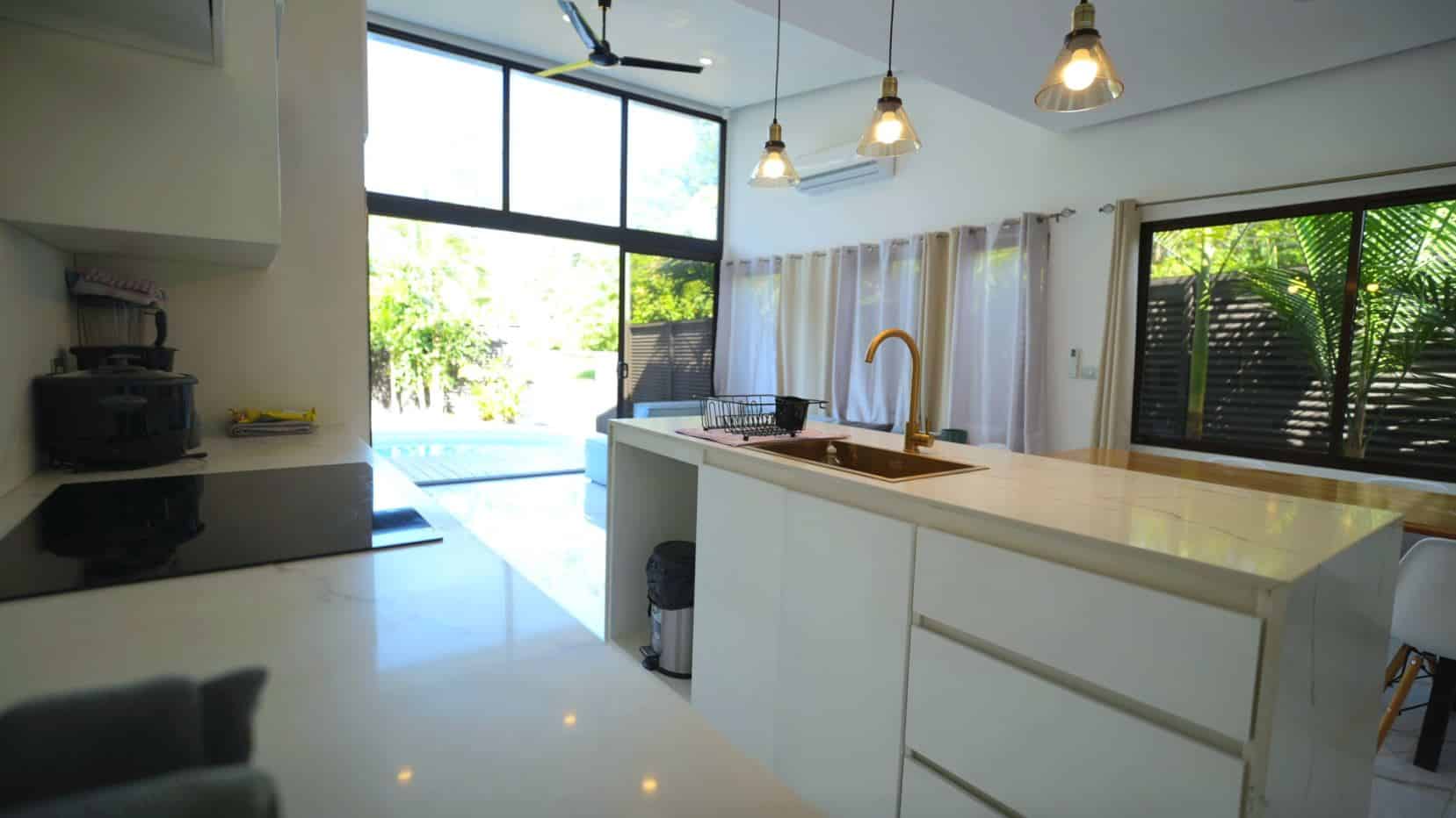 Bright, modern kitchen with white cabinetry and marble island, featuring a gold faucet and large windows overlooking a sunny outdoor space.