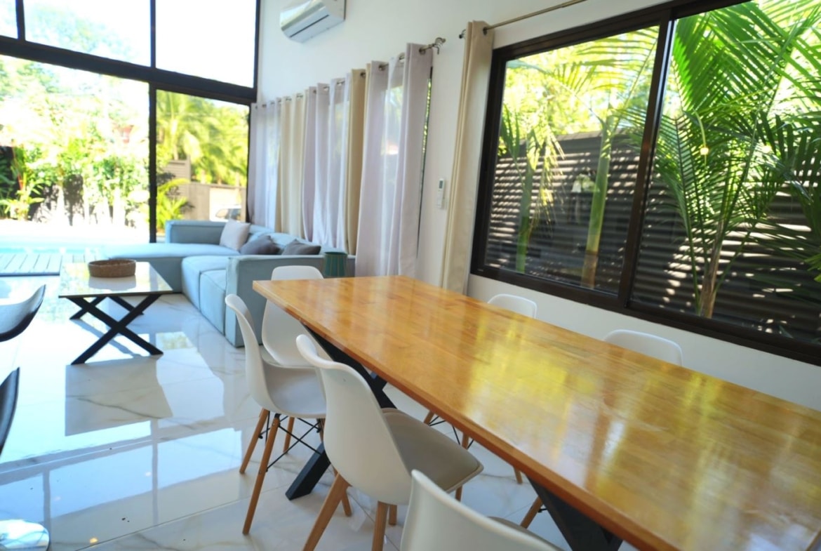 Bright open-plan living and dining area with a long wooden table, white chairs, a blue-gray sofa, and large glass windows showing greenery outside.
