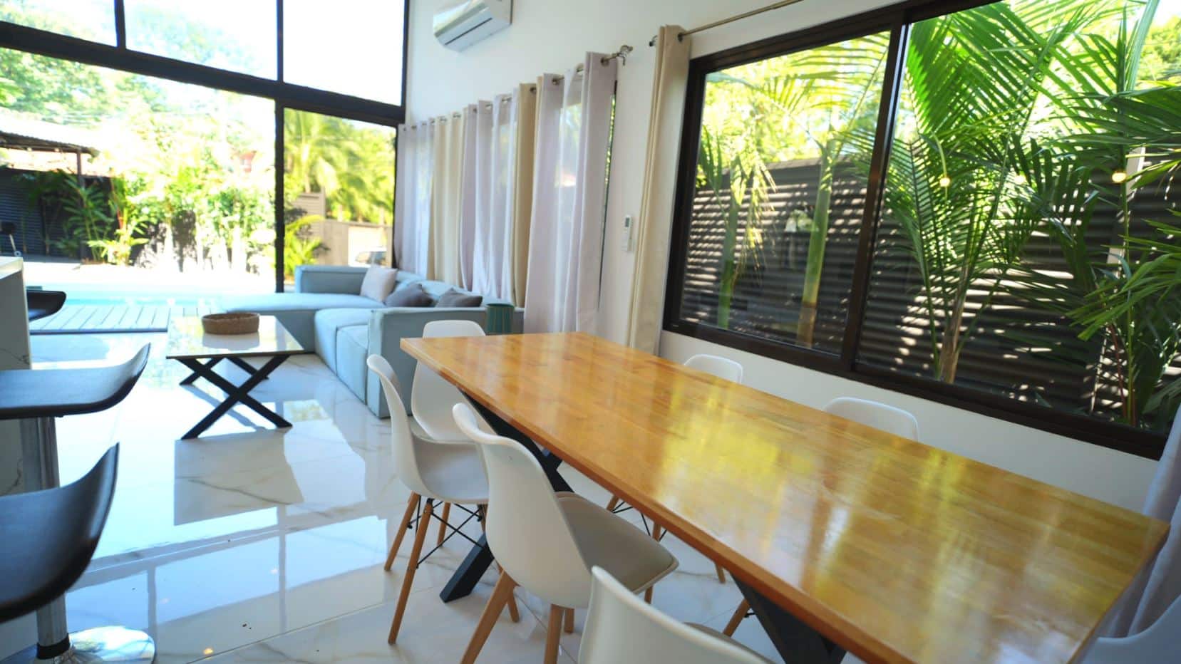 Bright open-plan living and dining area with a long wooden table, white chairs, a blue-gray sofa, and large glass windows showing greenery outside.