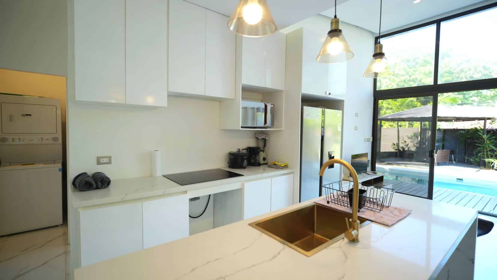 Bright modern kitchen with white cabinets, marble countertops, and a gold-finished sink overlooking a sunny outdoor pool through large glass doors.