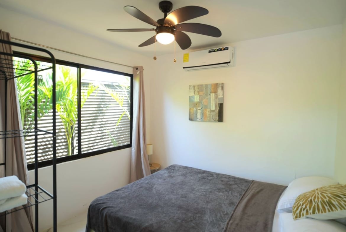 Bedroom with a large window, ceiling fan, and mounted air conditioner; bed in foreground with gray blanket and white/yellow pillows