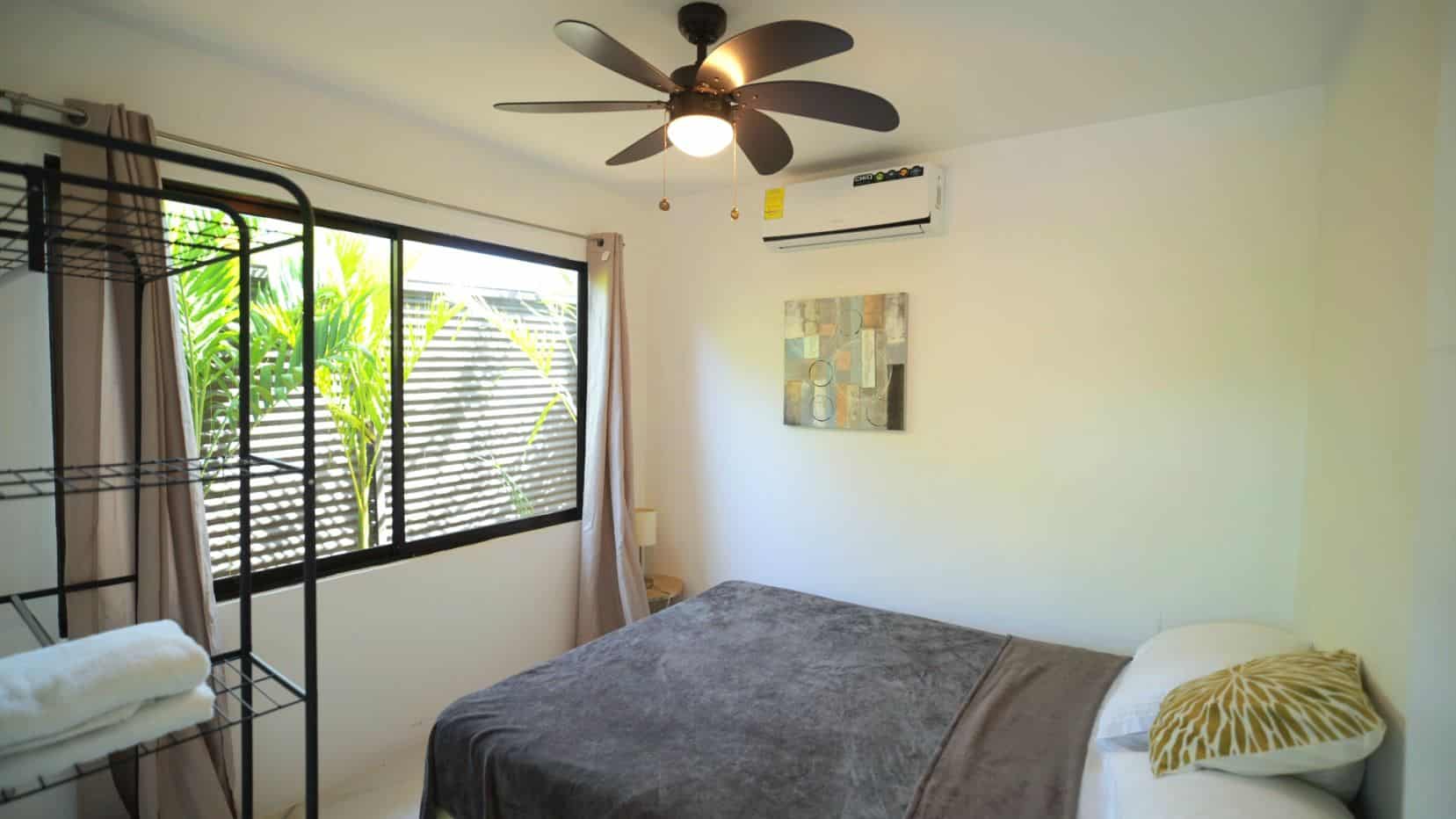 Bedroom with a large window, ceiling fan, and mounted air conditioner; bed in foreground with gray blanket and white/yellow pillows