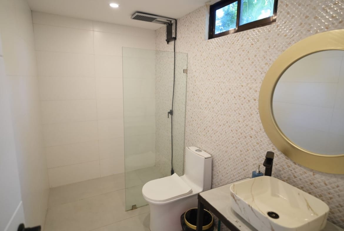 Modern bathroom with a glass shower enclosure, mosaic wall accent, toilet, and a marble sink with a large round beige-framed mirror above.