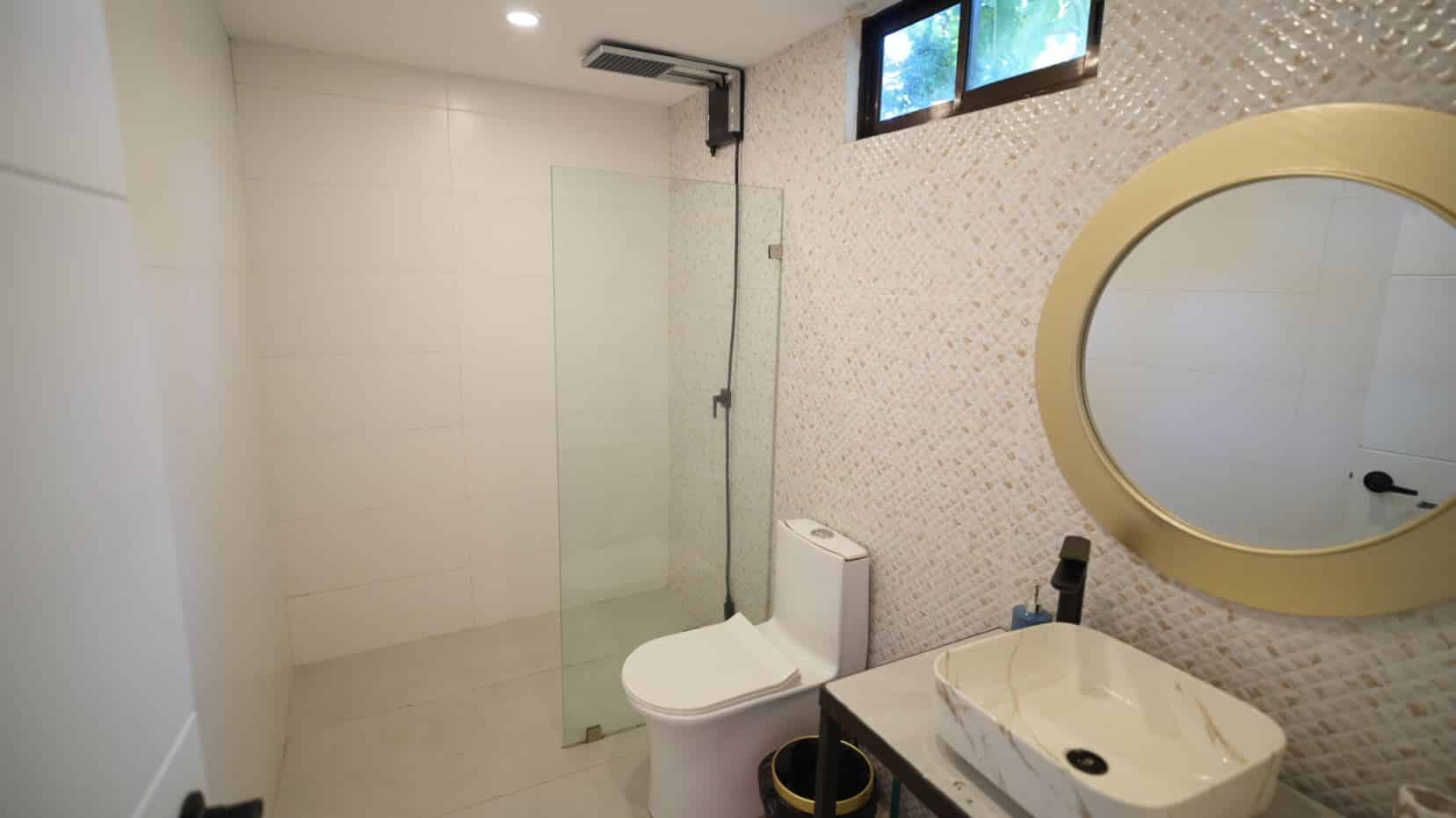 Modern bathroom with a glass shower enclosure, mosaic wall accent, toilet, and a marble sink with a large round beige-framed mirror above.