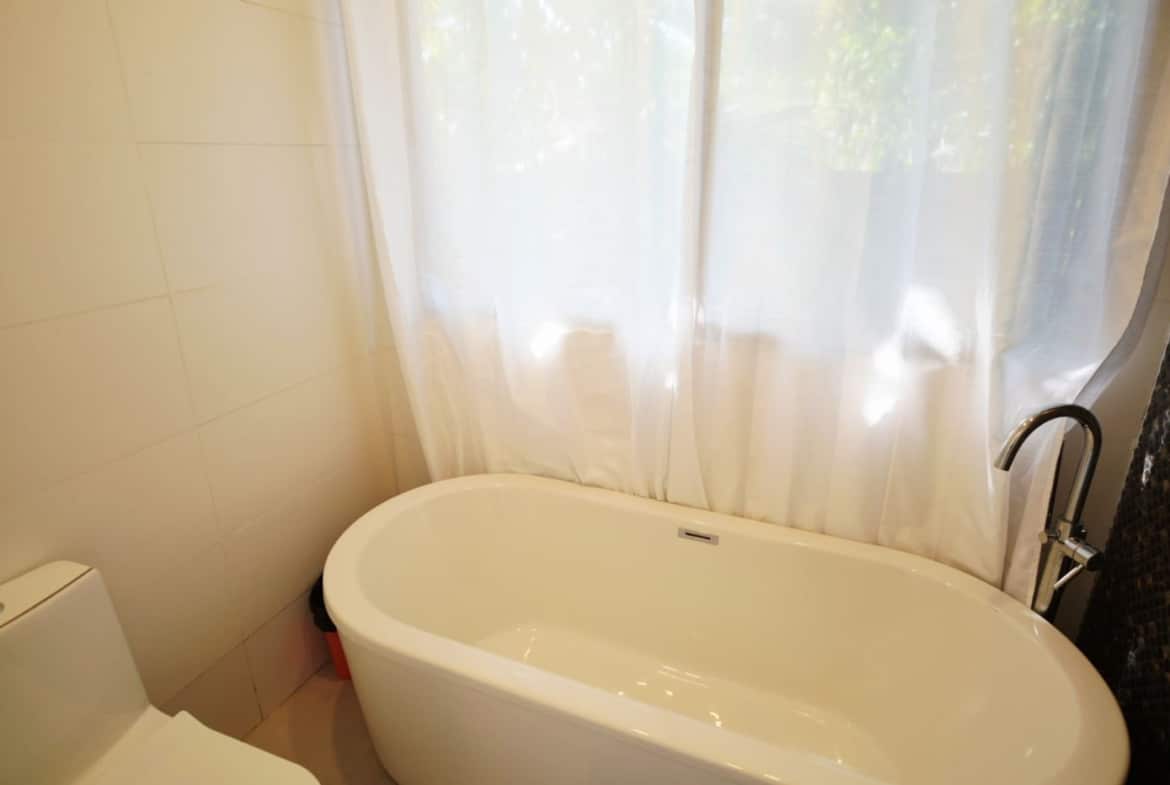 Freestanding white bathtub beside a window with sheer white curtains, chrome floor faucet, and tiled walls in a modern bathroom.