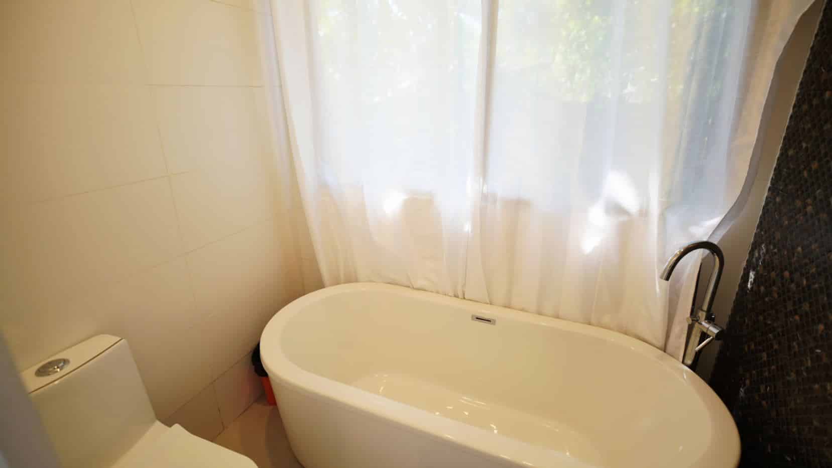 Freestanding white bathtub beside a window with sheer white curtains, chrome floor faucet, and tiled walls in a modern bathroom.