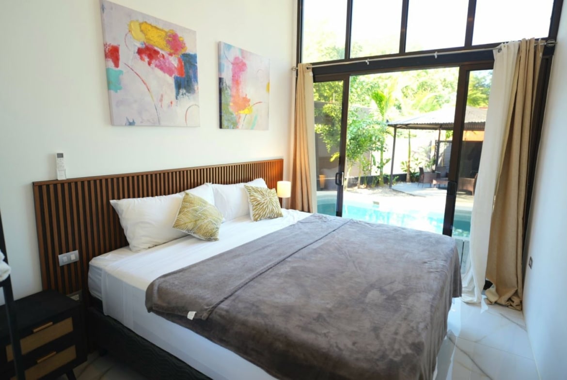King bed with white linens and gray throw, dark wood slatted headboard, two decorative pillows, nightstand and lamp nearby as abstract art hangs above.