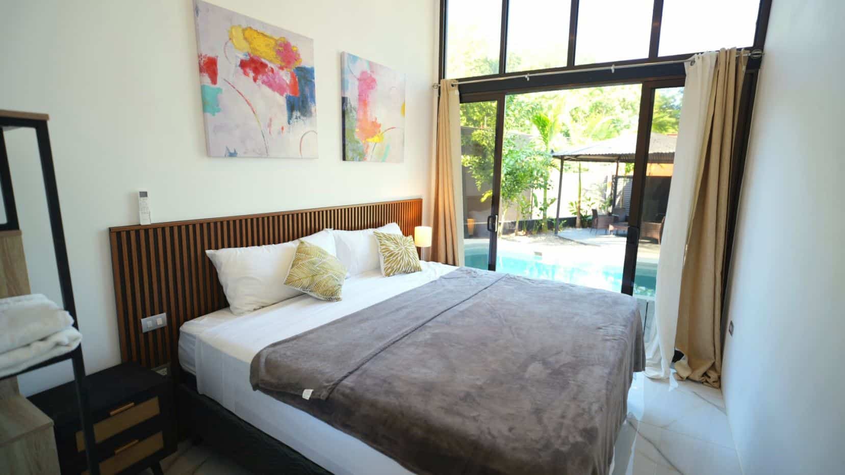 King bed with white linens and gray throw, dark wood slatted headboard, two decorative pillows, nightstand and lamp nearby as abstract art hangs above.