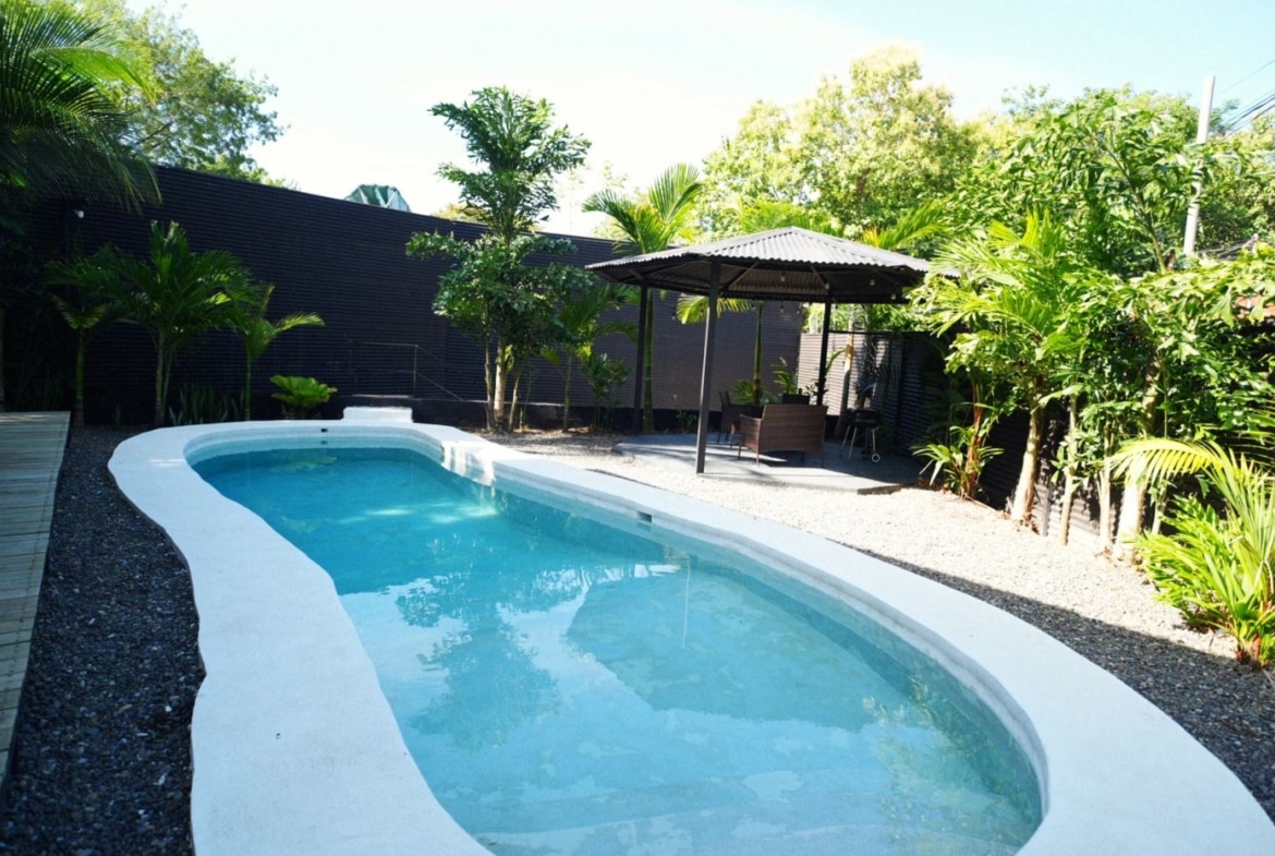 Backyard pool with a curved white edge, clear blue water, a wooden deck on the left, and a shaded seating area under a gazebo among tropical plants.