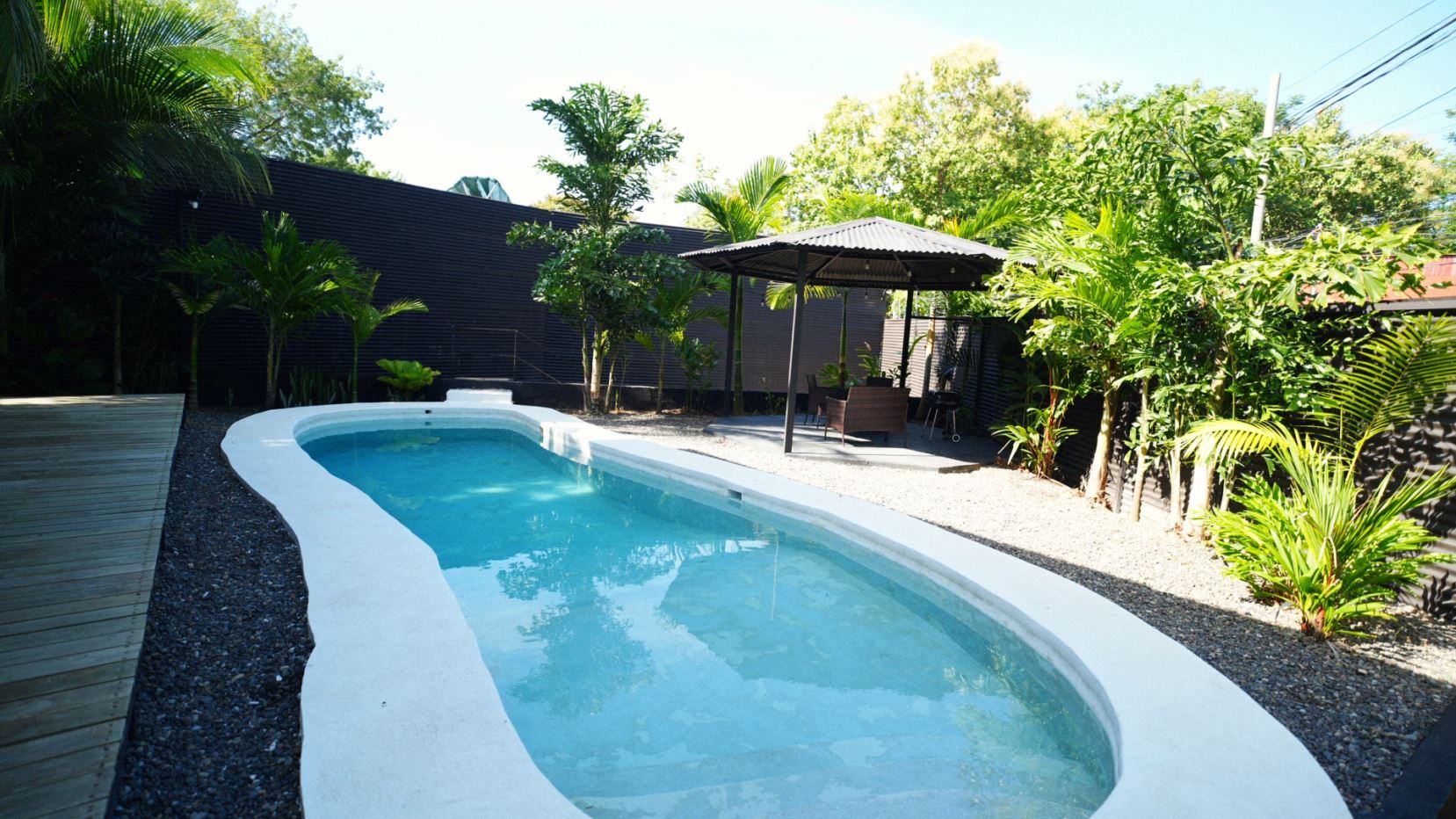 Backyard pool with a curved white edge, clear blue water, a wooden deck on the left, and a shaded seating area under a gazebo among tropical plants.