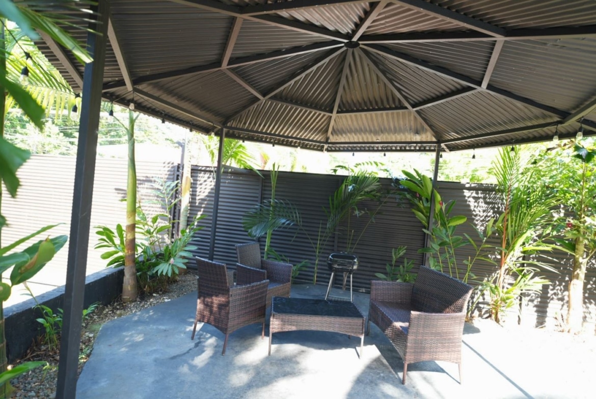 Outdoor gazebo-covered patio with brown wicker chairs around a glass coffee table and lush tropical plants along a corrugated fence.