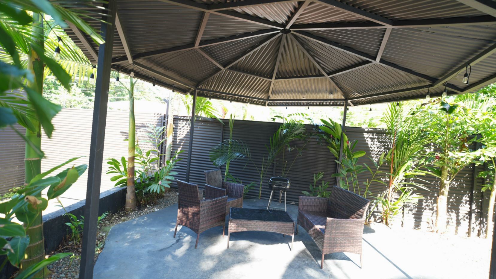 Outdoor gazebo-covered patio with brown wicker chairs around a glass coffee table and lush tropical plants along a corrugated fence.