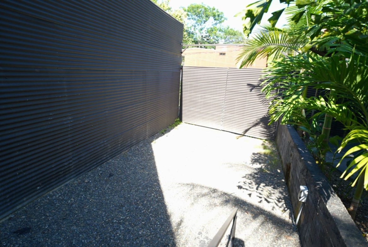 Narrow gravel alley between dark corrugated metal fences, ending at a metal gate, with tropical plants along the right.