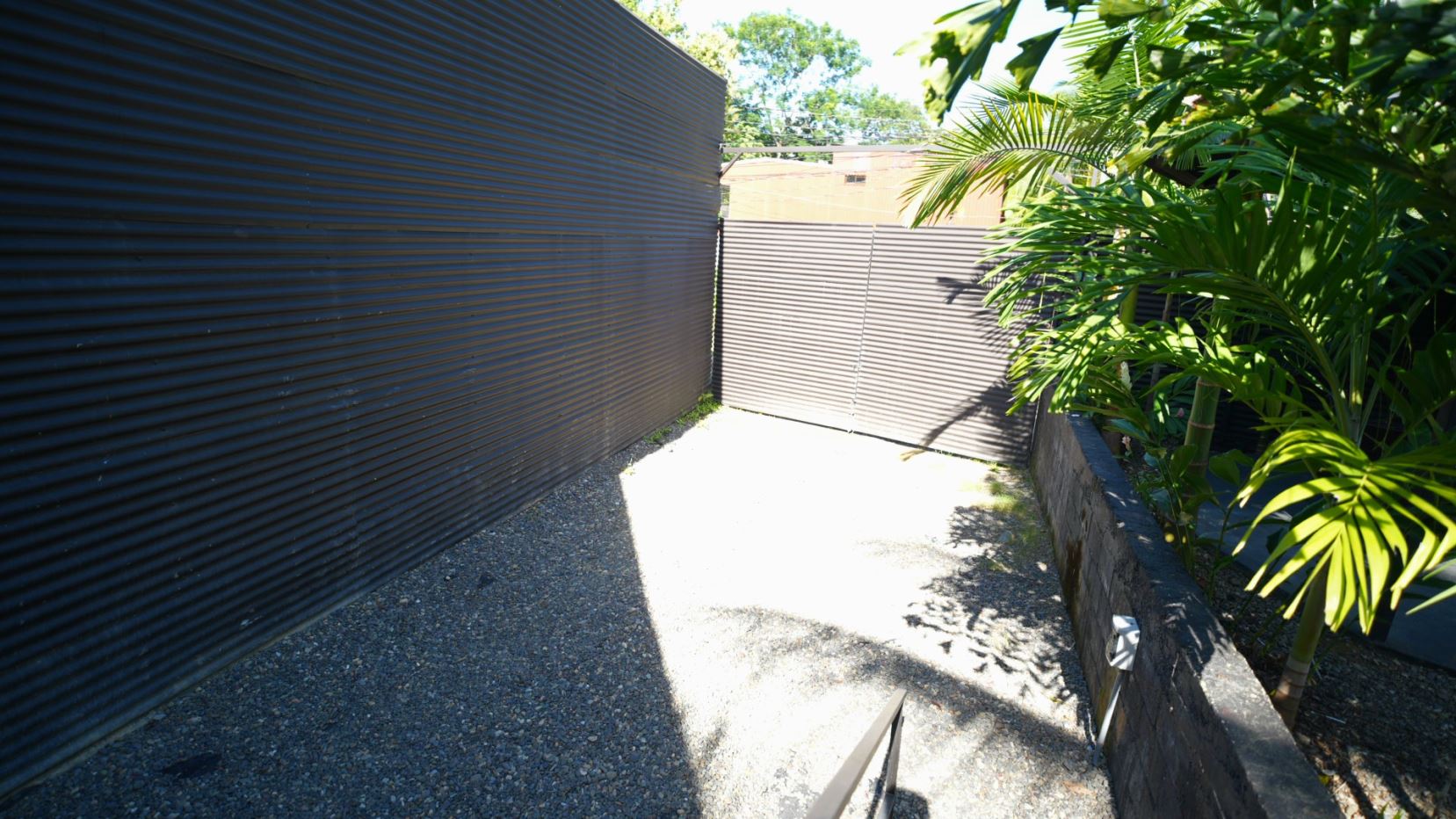 Narrow gravel alley between dark corrugated metal fences, ending at a metal gate, with tropical plants along the right.