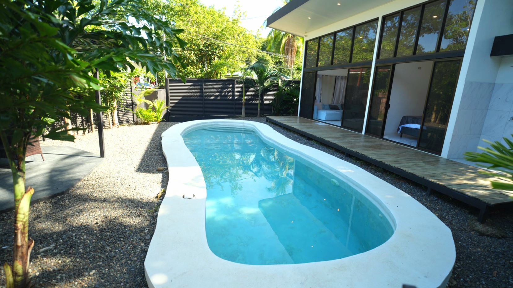 Kidney-shaped pool beside a modern glass-walled house with a wooden deck and tropical plants nearby.