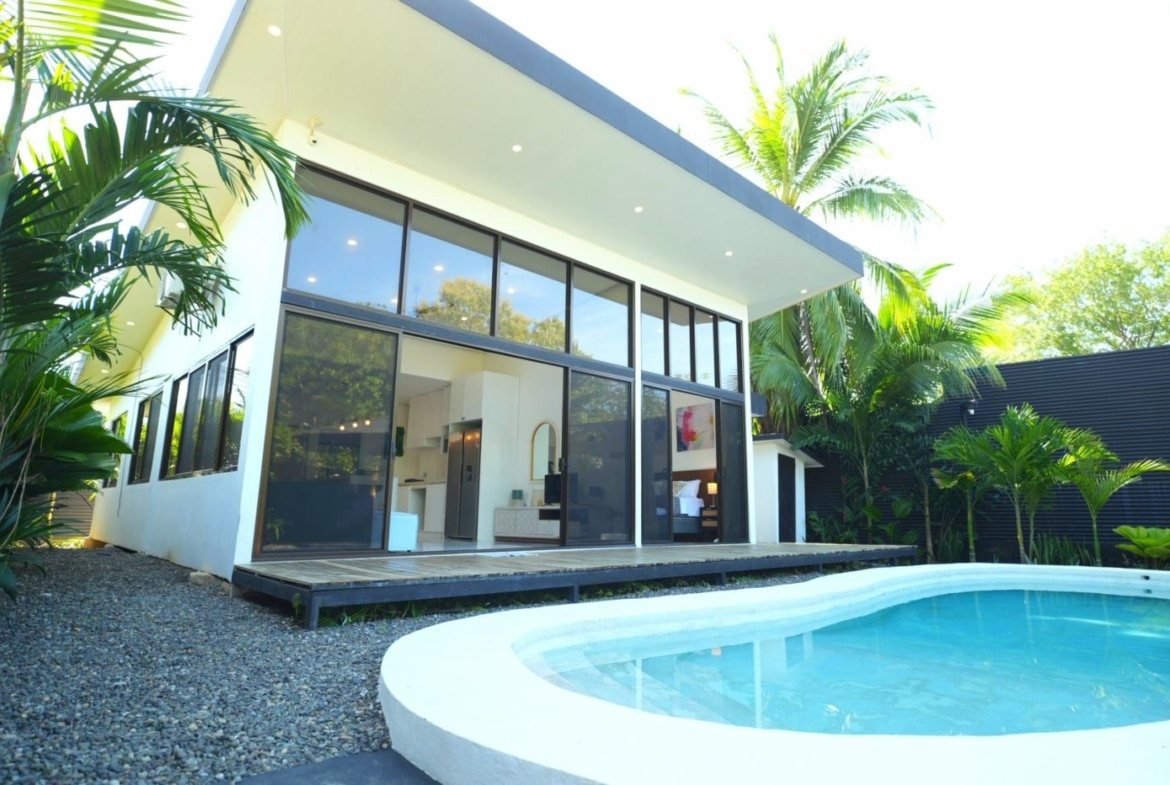 Modern white house with floor-to-ceiling glass windows beside a curved outdoor pool and tropical plants.
