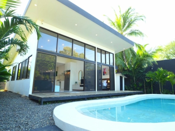 Modern white house with floor-to-ceiling glass windows beside a curved outdoor pool and tropical plants.