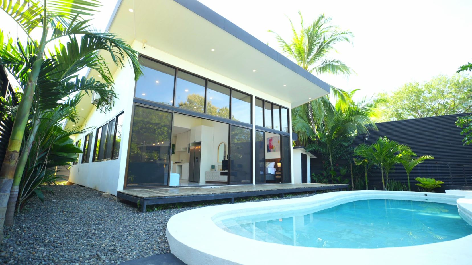 Modern white house with floor-to-ceiling glass windows beside a curved outdoor pool and tropical plants.