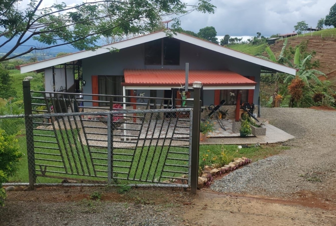 Gate leads to a blue single-story house with a red roof amid a green hillside yard, trees overhead. (informational)