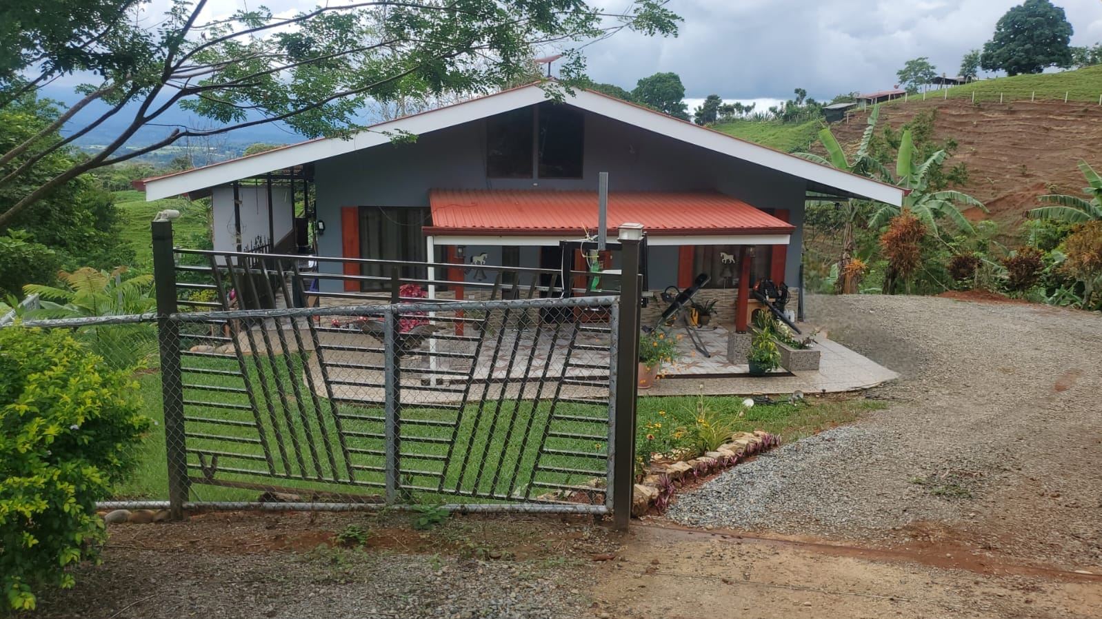 Gate leads to a blue single-story house with a red roof amid a green hillside yard, trees overhead. (informational)