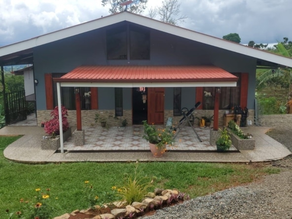 Single-story blue-gray house with a red metal roof over a covered porch, surrounded by a gravel driveway and lush garden plants.