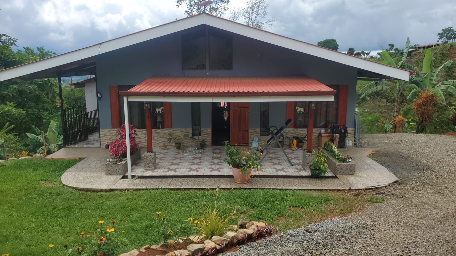 Single-story blue-gray house with a red metal roof over a covered porch, surrounded by a gravel driveway and lush garden plants.