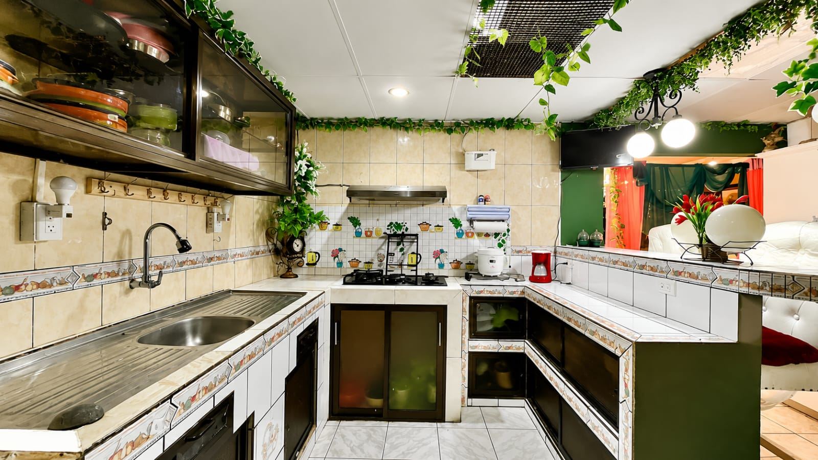 U-shaped kitchen with tiled counters, sink, stove, and hanging greenery along the ceiling.