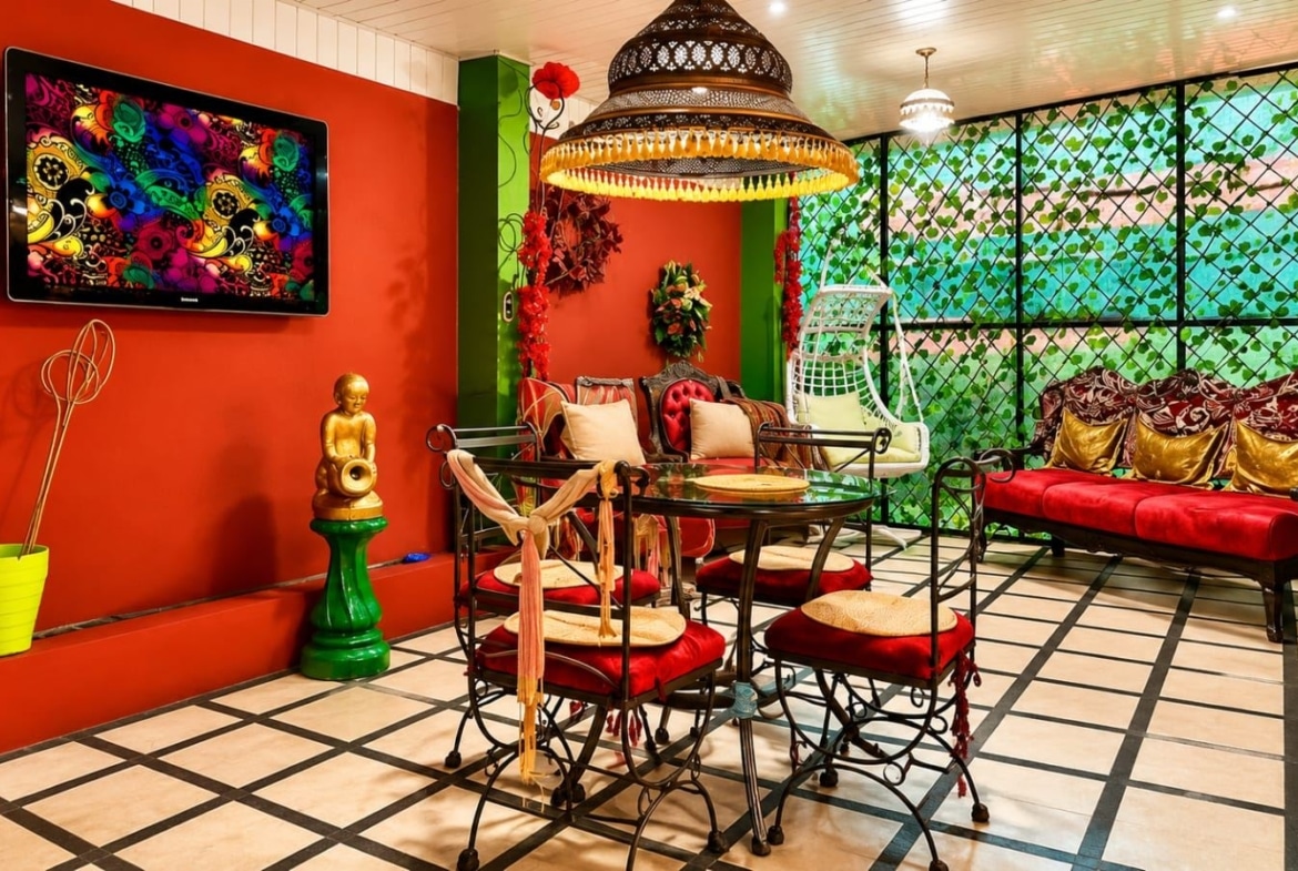 Vibrant indoor lounge with red walls, wrought-iron furniture, red cushions, and a colorful wall TV art piece on the left.
