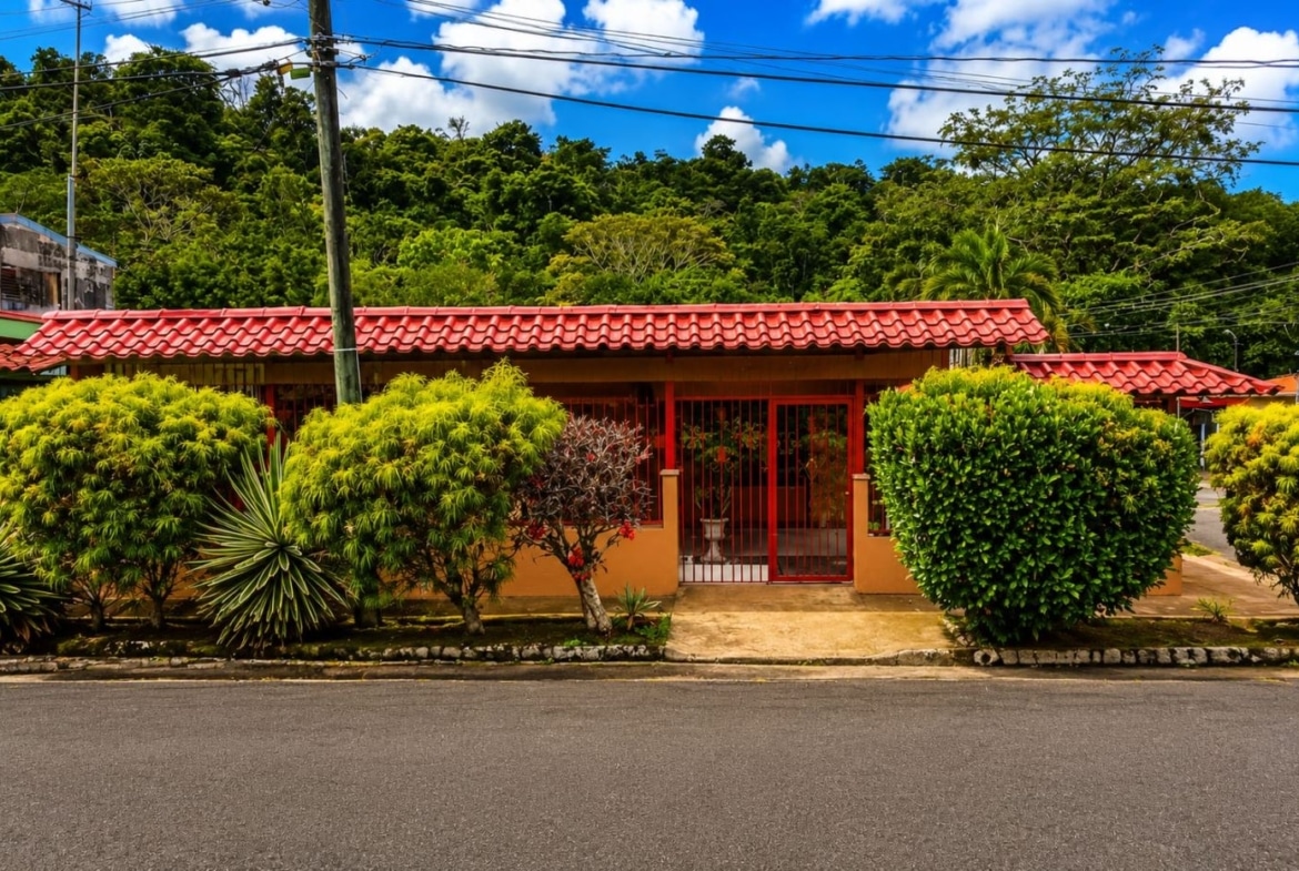 Red-tiled, orange-walled house with a red gate, flanked by lush tropical shrubs on a residential street. Sky is blue with clouds above.
