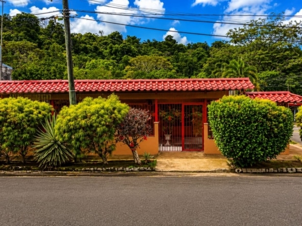 Red-tiled, orange-walled house with a red gate, flanked by lush tropical shrubs on a residential street. Sky is blue with clouds above.