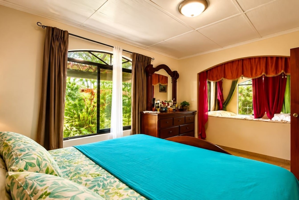 Bright hotel bedroom with a turquoise bedspread, floral pillows, and a large window overlooking lush greenery outside.