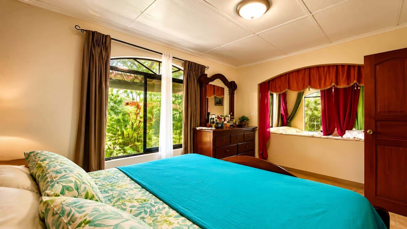 Bright hotel bedroom with a turquoise bedspread, floral pillows, and a large window overlooking lush greenery outside.