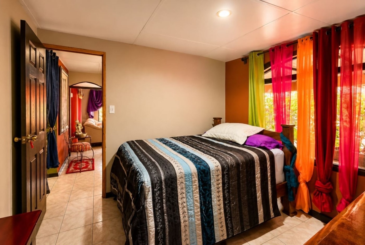 Bedroom with a striped quilt on a wooden bed, bright multicolored curtains by the window, and tiled floor.
