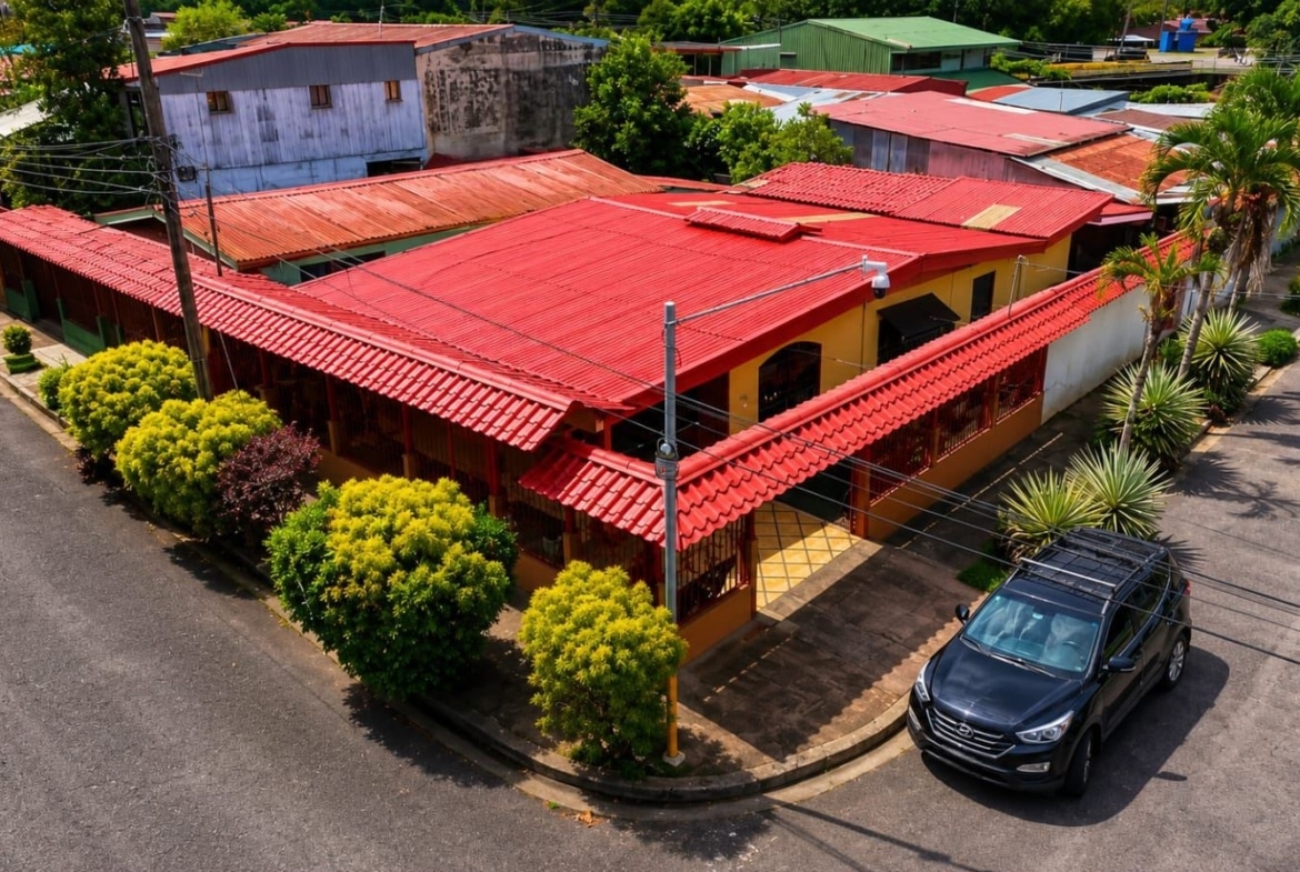 Aerial view of a yellow building with a bright red roof at a street corner, surrounded by green shrubs and a dark SUV parked nearby.