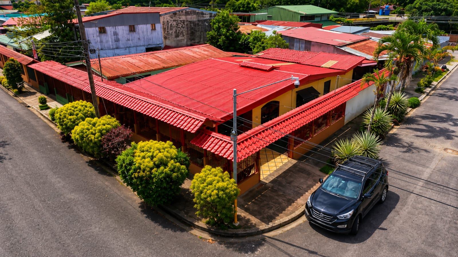 Aerial view of a yellow building with a bright red roof at a street corner, surrounded by green shrubs and a dark SUV parked nearby.