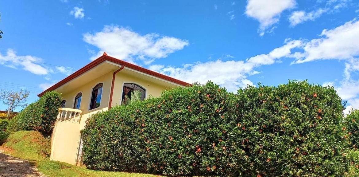 Single-story yellow house with a red roof partially hidden by a tall green hedge on a sunny day. Arched windows overlook a small balcony.