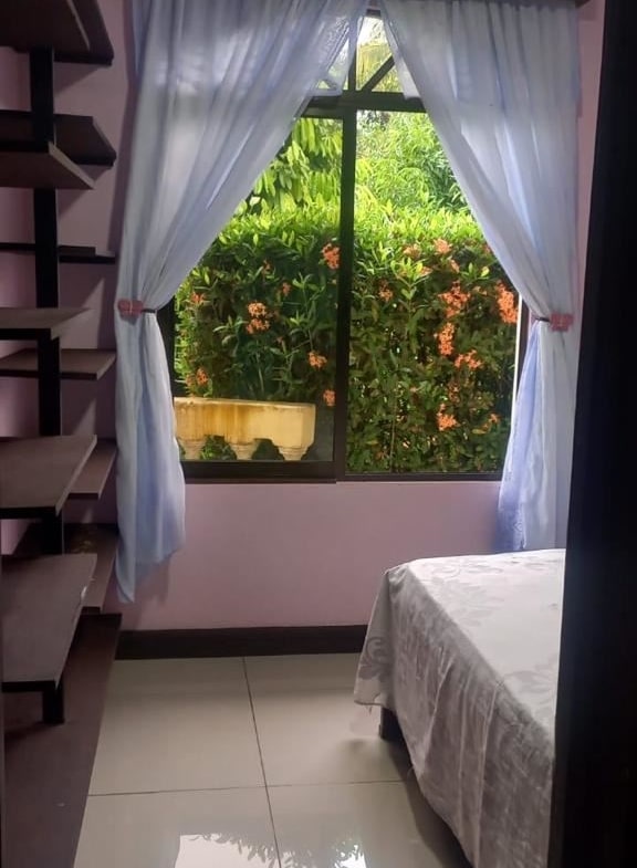 Bedroom with pink walls, white curtains tied back, window showing green bushes and orange flowers outside, and a bed with a light patterned cover on the right.